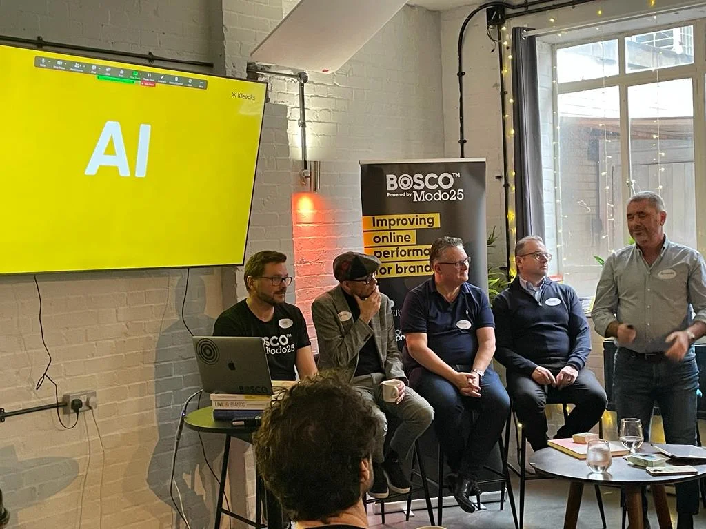 Panel of five speakers discusses AI at a Bosco Modo25 event, with a yellow "AI" slide displayed on screen.