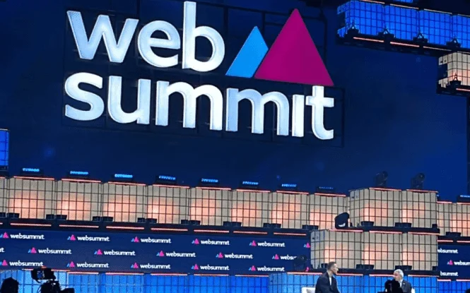 Two speakers seated on stage at Web Summit in front of a large illuminated logo backdrop.