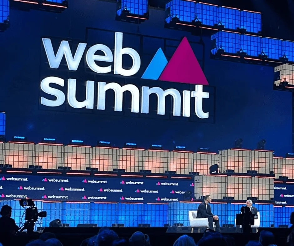 Two speakers seated on stage at Web Summit in front of a large illuminated logo backdrop.
