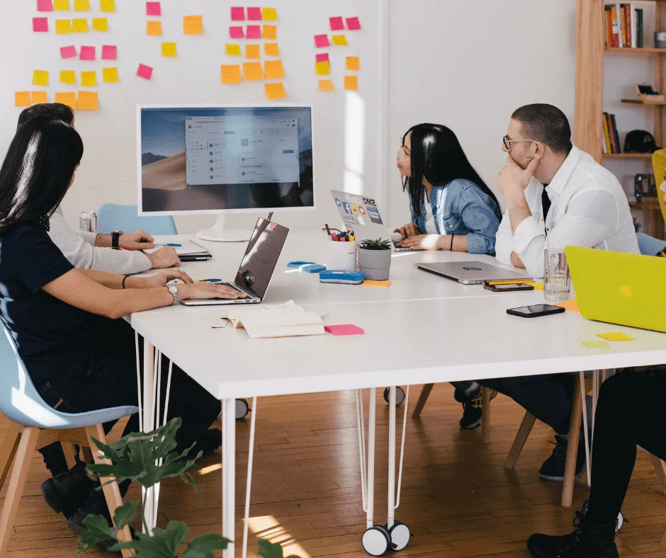 Team collaborating around a laptop-filled table during a market research business strategy meeting with sticky notes on the wall. Team collaborating around a laptop-filled table during a market research business strategy meeting with sticky notes on the wall.