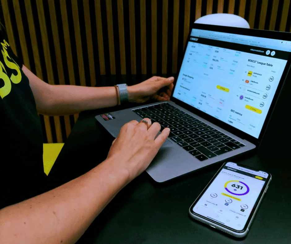 Person analyzing BOSCO dashboard on laptop and phone, showing competitor insights and SEO performance metrics.