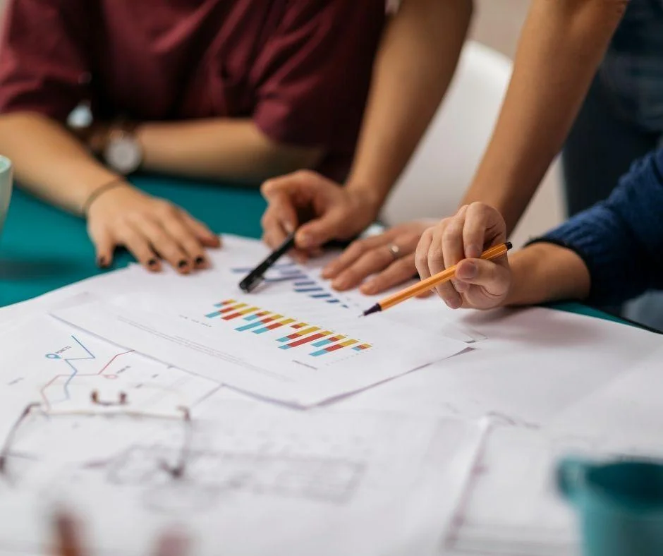 Three people collaborate over bar chart data and blueprints, discussing forecasting trends at a table.