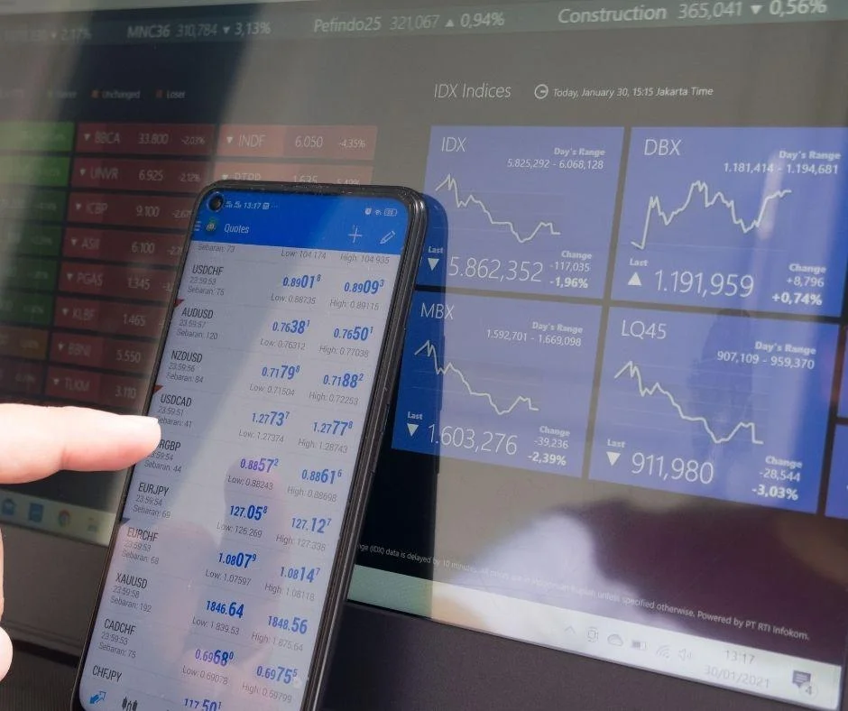 A smartphone displaying forex quotes held up to a screen with stock indices, illustrating data used in predictive analytics models.