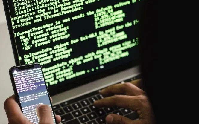 Global click fraud detection concept showing green code on a laptop screen while a hand checks a smartphone.