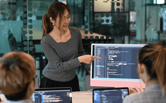 Woman explains code on a monitor to colleagues, illustrating data-driven eCommerce marketing strategies.