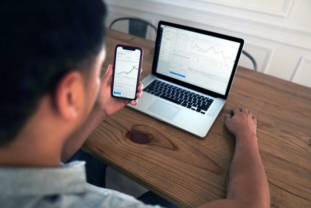 Man reviewing stock charts on phone and laptop, analyzing historical universal analytics data trends.