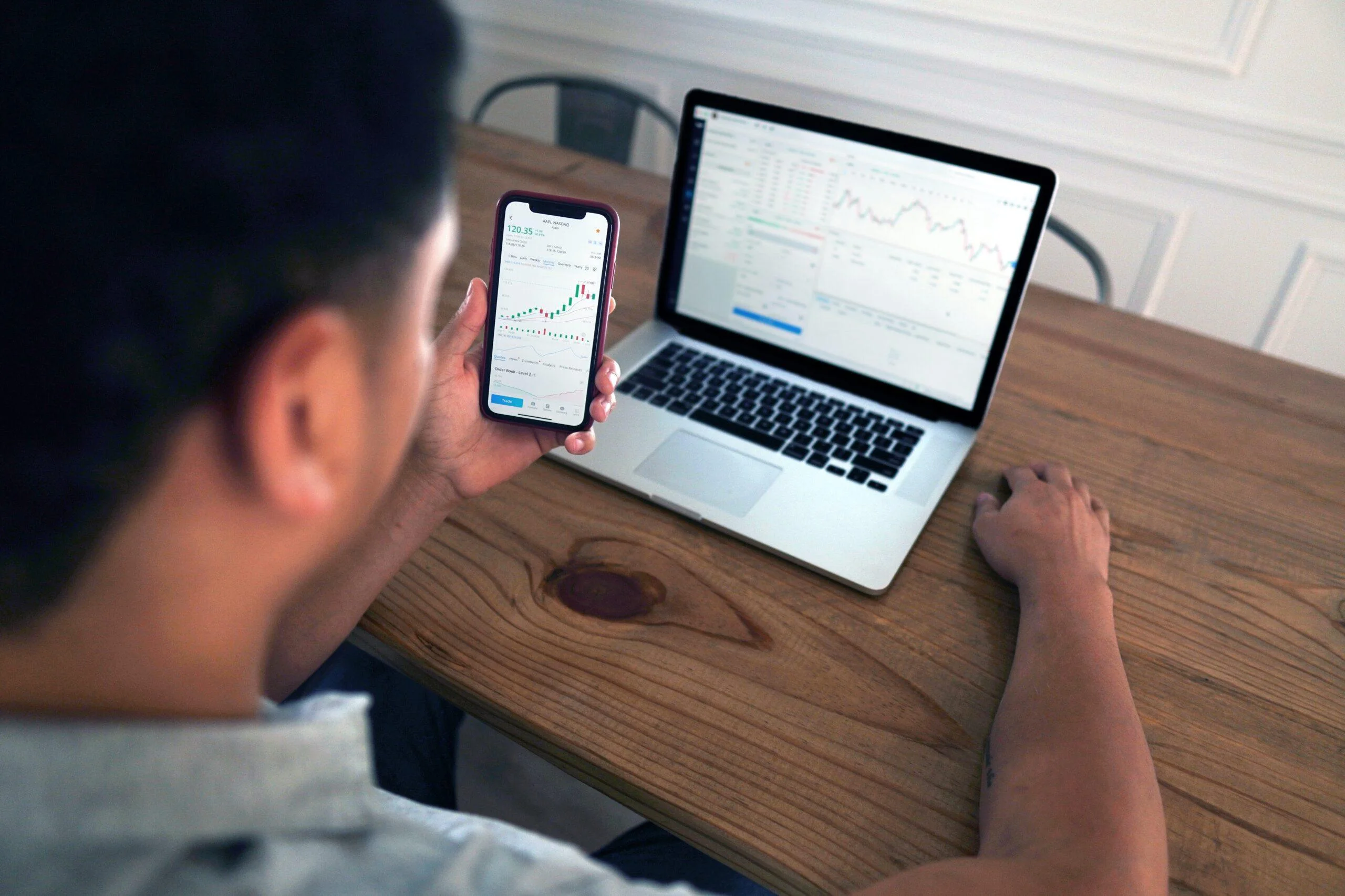 Man reviewing stock charts on phone and laptop, analyzing historical universal analytics data trends.